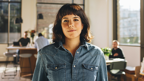 Osoba stoi w biurze, ubrana w niebieską koszulę dżinsową. W tle widać innych pracowników przy stolikach, co sugeruje kreatywną atmosferę sprzyjającą pracy i spotkaniom biznesowym.