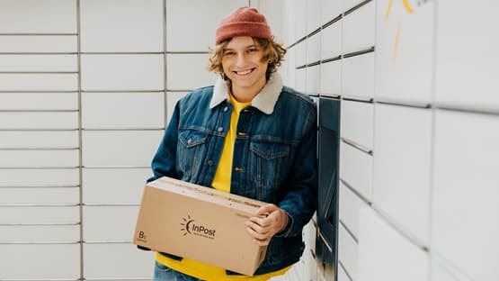 Osoba trzymająca paczkę InPost przy maszynie Paczkomat w kurtce jeansowej i czerwonej czapce.