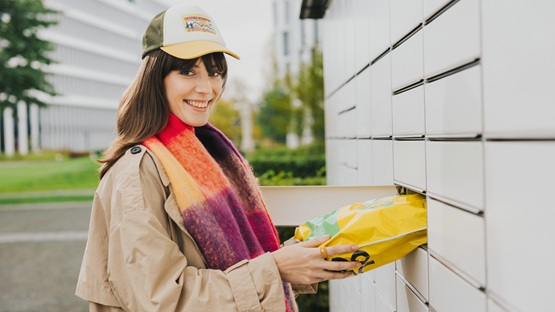 Uśmiechnięta osoba w płaszczu i szaliku umieszcza paczkę w skrytce maszyny Paczkomat, gotowa do nadania przesyłki za granicę.