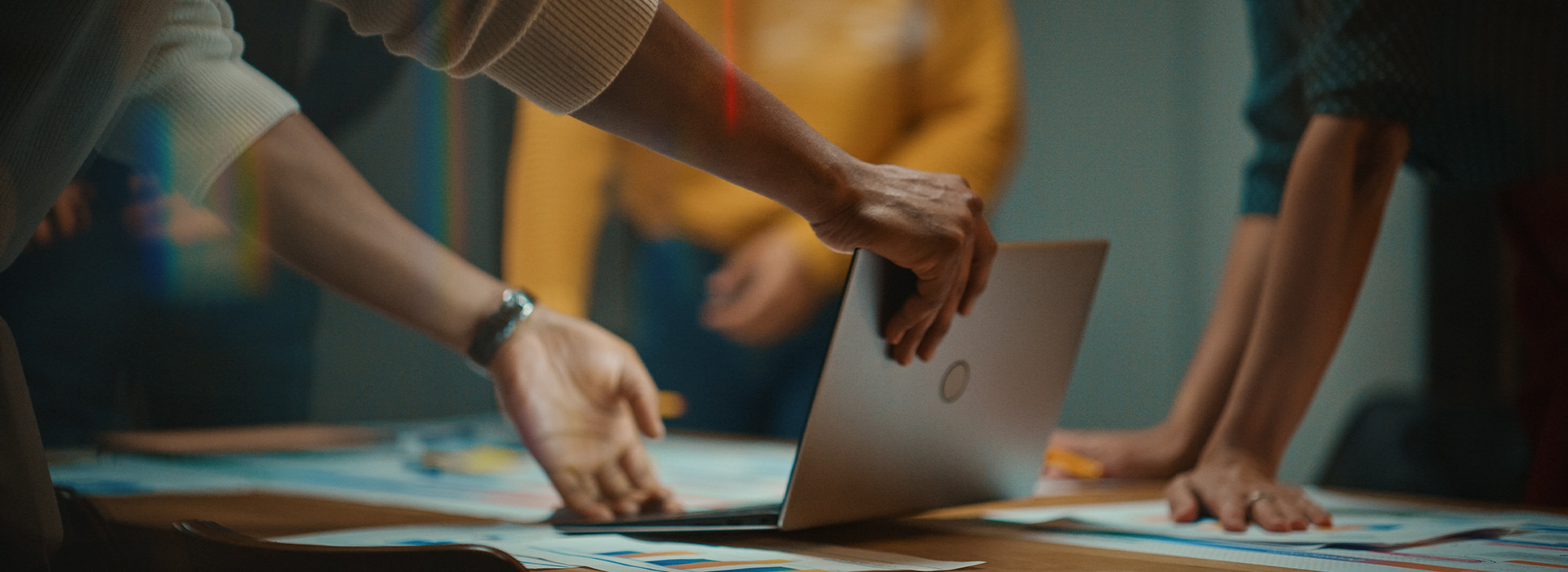 Ręce ludzi przy biurku nad laptopem i dokumentami, podczas spotkania o automatyzacji sprzedaży przez internet.