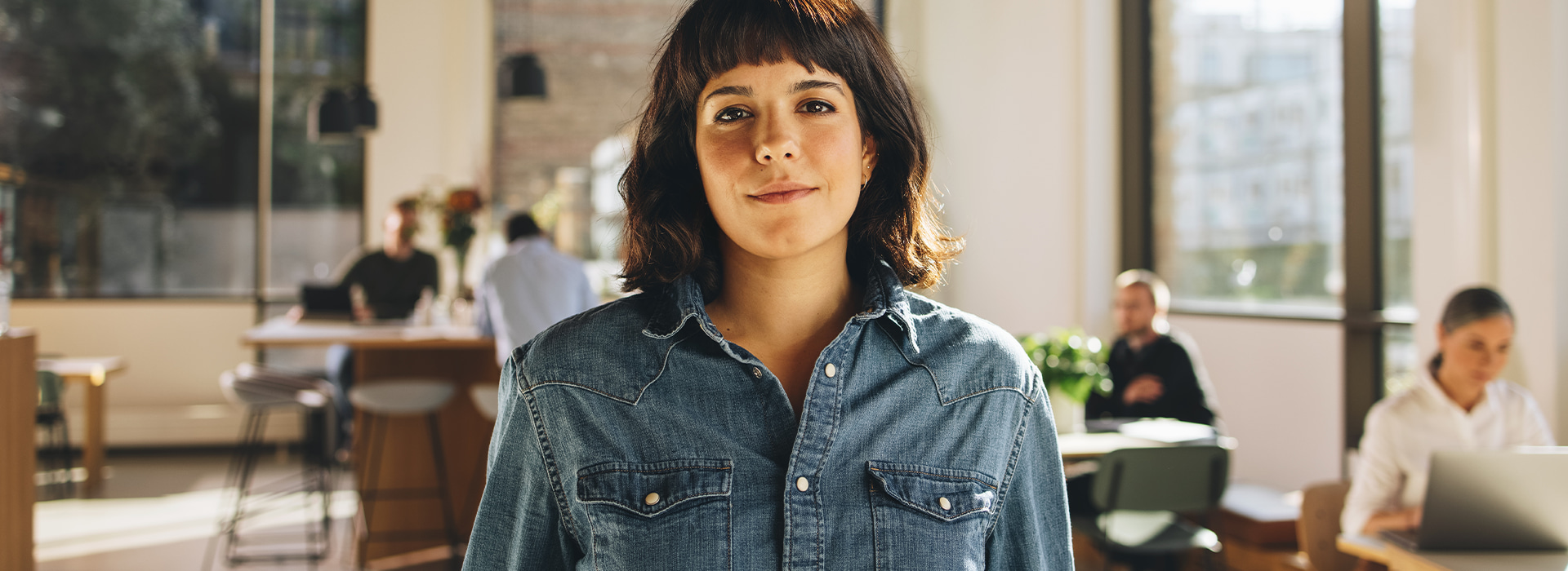 Osoba stoi w biurze, ubrana w niebieską koszulę dżinsową. W tle widać innych pracowników przy stolikach, co sugeruje kreatywną atmosferę sprzyjającą pracy i spotkaniom biznesowym.