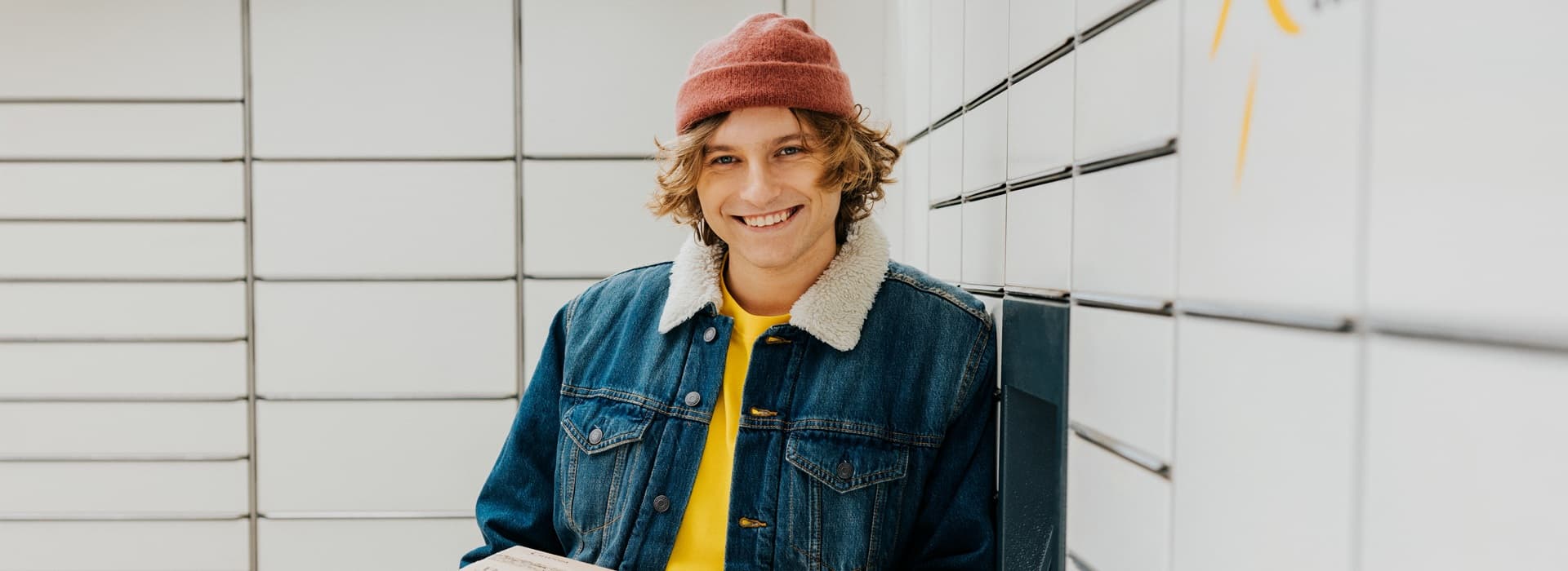 Osoba trzymająca paczkę InPost przy maszynie Paczkomat w kurtce jeansowej i czerwonej czapce.