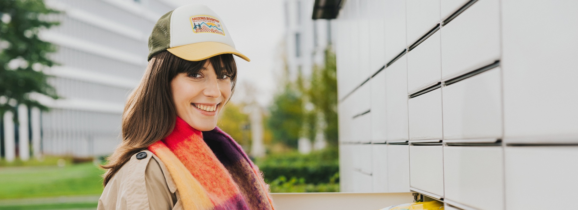 Uśmiechnięta osoba w płaszczu i szaliku umieszcza paczkę w skrytce maszyny Paczkomat, gotowa do nadania przesyłki za granicę.