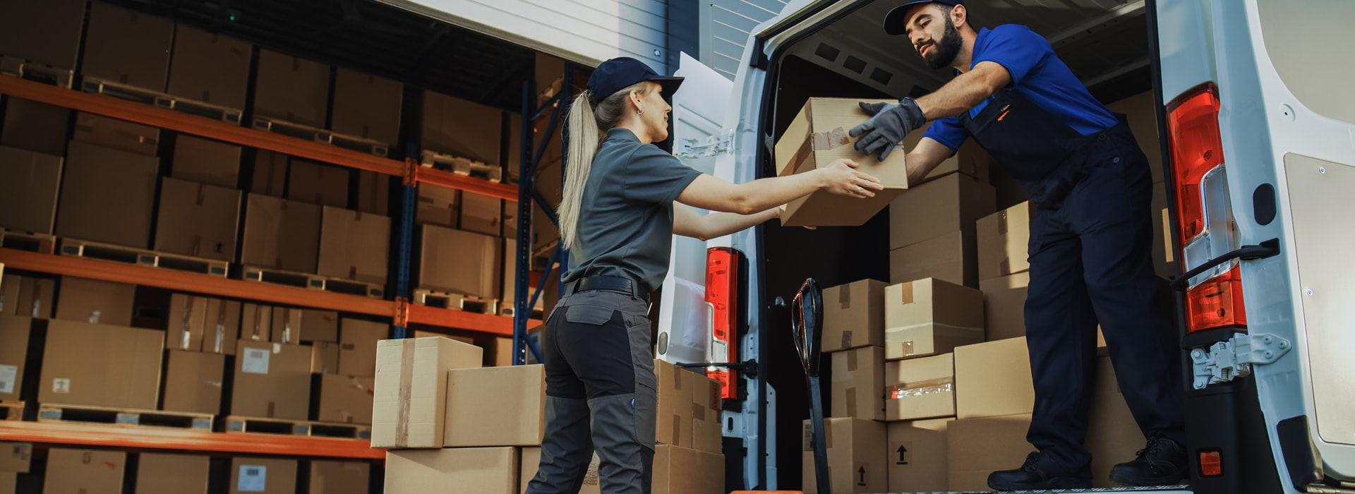 Transport drobnicowy: kurier przekazuje paczkę współpracowniczce przy otwartym samochodzie dostawczym, w tle regały magazynowe z kartonami.