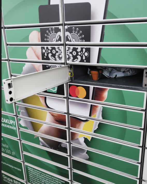Otwarta skrytka urządzenia Paczkomat w okleinie kampanii Banków Spółdzielczych z Grupy BPS i Mastercard. W środku niezapominajka oraz paczka.