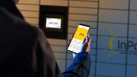 Osoba stoi przez maszyną Paczkomat z wyciągniętym telefonem, na ekranie którego jest otwarta aplikacja InPost Mobile.