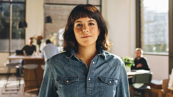 Osoba stoi w biurze, ubrana w niebieską koszulę dżinsową. W tle widać innych pracowników przy stolikach, co sugeruje kreatywną atmosferę sprzyjającą pracy i spotkaniom biznesowym.