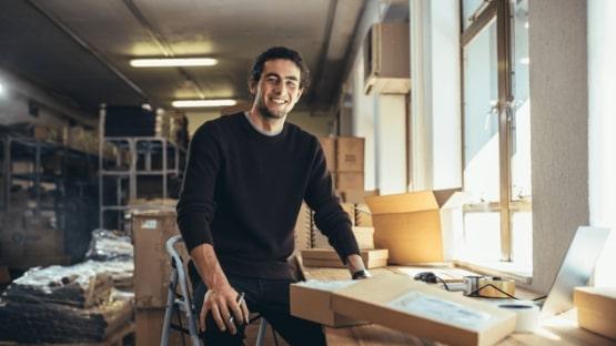 Osoba siedzi w magazynie otoczona kartonami i materiałami do pakowania. Ubrana jest w ciemny sweter, a obok znajduje się biurko z komputerem. Przestrzeń jest dobrze oświetlona, a w tle są półki.