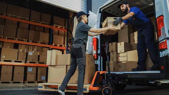 Transport drobnicowy: kurier przekazuje paczkę współpracowniczce przy otwartym samochodzie dostawczym, w tle regały magazynowe z kartonami.
